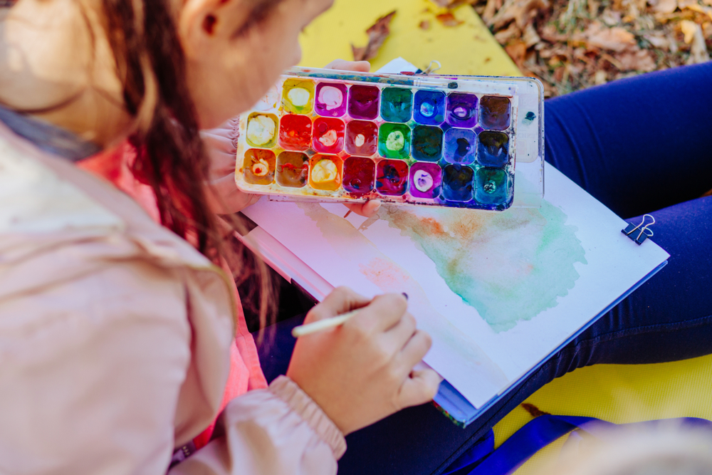 Young girl painting with watercolour paint on white paper on her lap